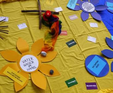 Quelle: ekiba_amd Bodenbild: Auf einem gelbenTuch loegen eine ausgeschnittene Blume mit Dem Text Vater unser im Himmel, ein Kreuz aus Holzstöcken, eine Kerze und verschieden Farbige Kärtchen